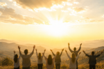 A group of people facing away from the camera with hands raised as the sun rises.