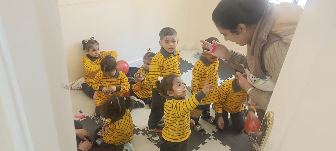 A group of young children with face paint, balloons, and yellow striped shirts playing in a small room of a school in Al-Arish, Egypt.