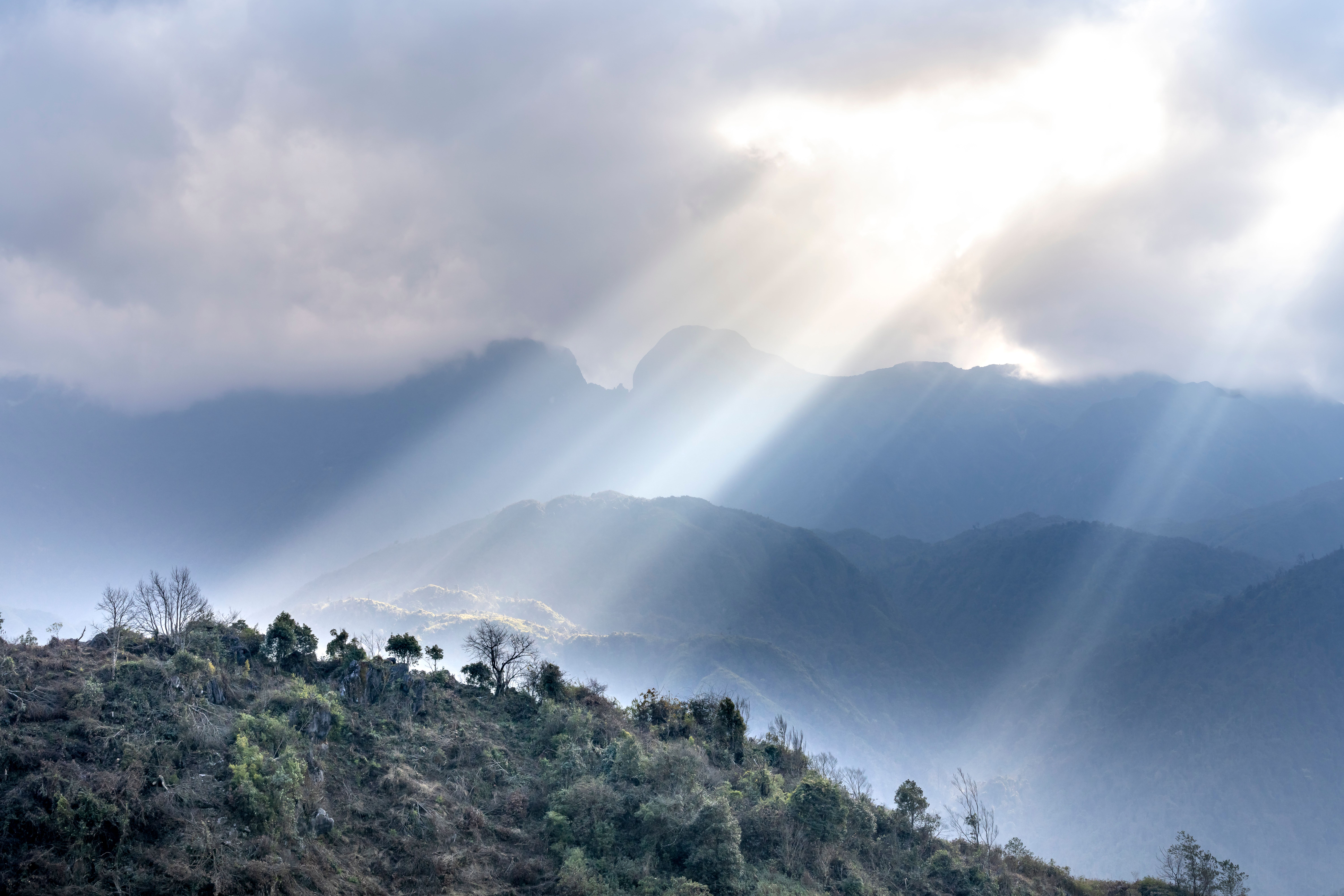 Green hill in the foreground and hills that look blue in the background. A strong sunbeam shines down through parted clouds in the sky.